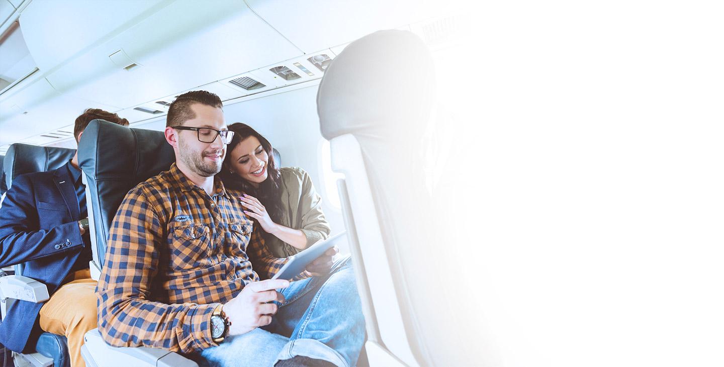 A man and woman sitting next to each other enjoying the passenger experience on an airplane, looking at a tablet